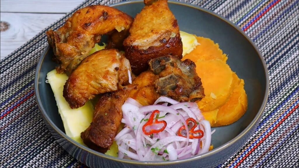 Trozos de cerdo dorados acompañados de yuca y camote sancochados, servidos con cebolla morada encurtida y ají, dispuestos en plato hondo sobre mantel artesanal colorido