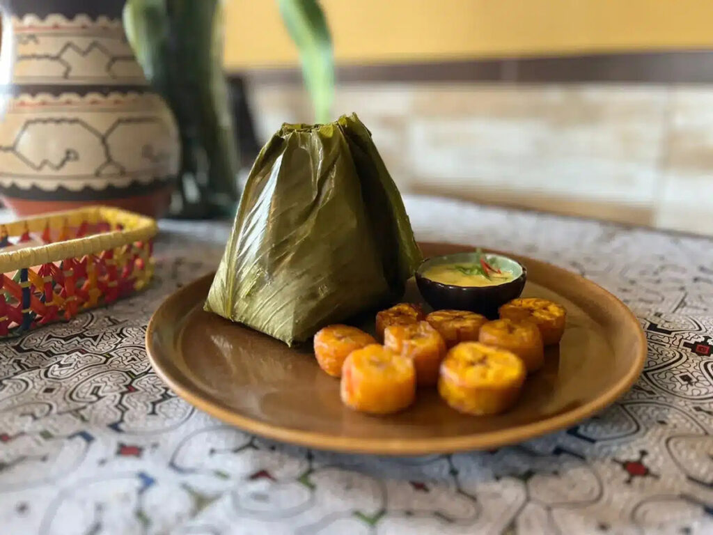 Juane de la selva envuelto en hojas de bijao, acompañado de plátanos fritos en rodajas y crema de ají, servido sobre plato de cerámica con mantel de diseño shipibo