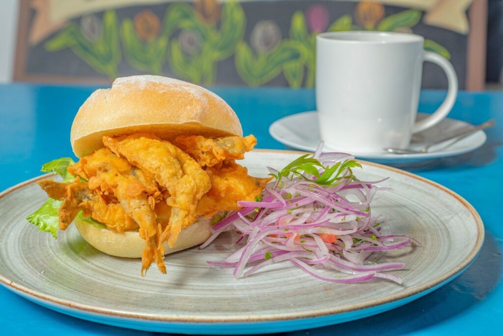 Pan francés relleno de pejerreyes fritos dorados, acompañado de cebolla morada en juliana y lechuga fresca, servido en plato de cerámica sobre mesa azul con taza de café al fondo
