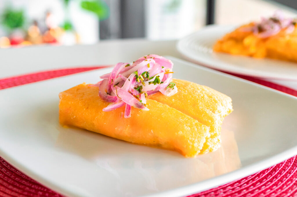 Tamal limeño envuelto en maíz amarillo, coronado con cebolla morada encurtida, presentado en plato blanco con mantel rojo, resaltando su textura suave y color dorado.