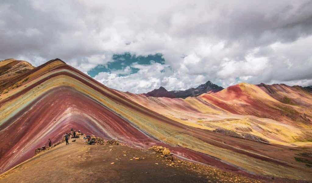 Vista panorámica de la montaña Vinicunca en Cusco, mostrando las franjas de colores rojo, amarillo y turquesa bajo un cielo andino nublado