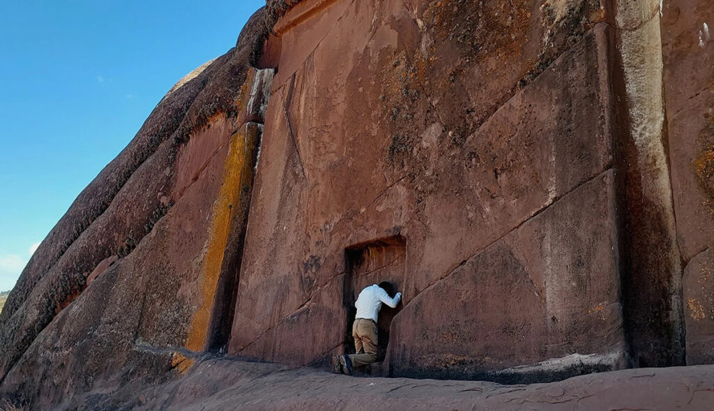 Visitante frente al portal de Aramu Muru en el altiplano, realizando una ofrenda o meditación junto a la roca sagrada