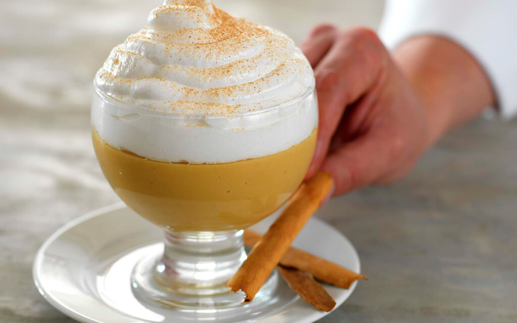 Copa de suspiro a la limeña con merengue espolvoreado con canela, servida con ramas de canela en un plato blanco