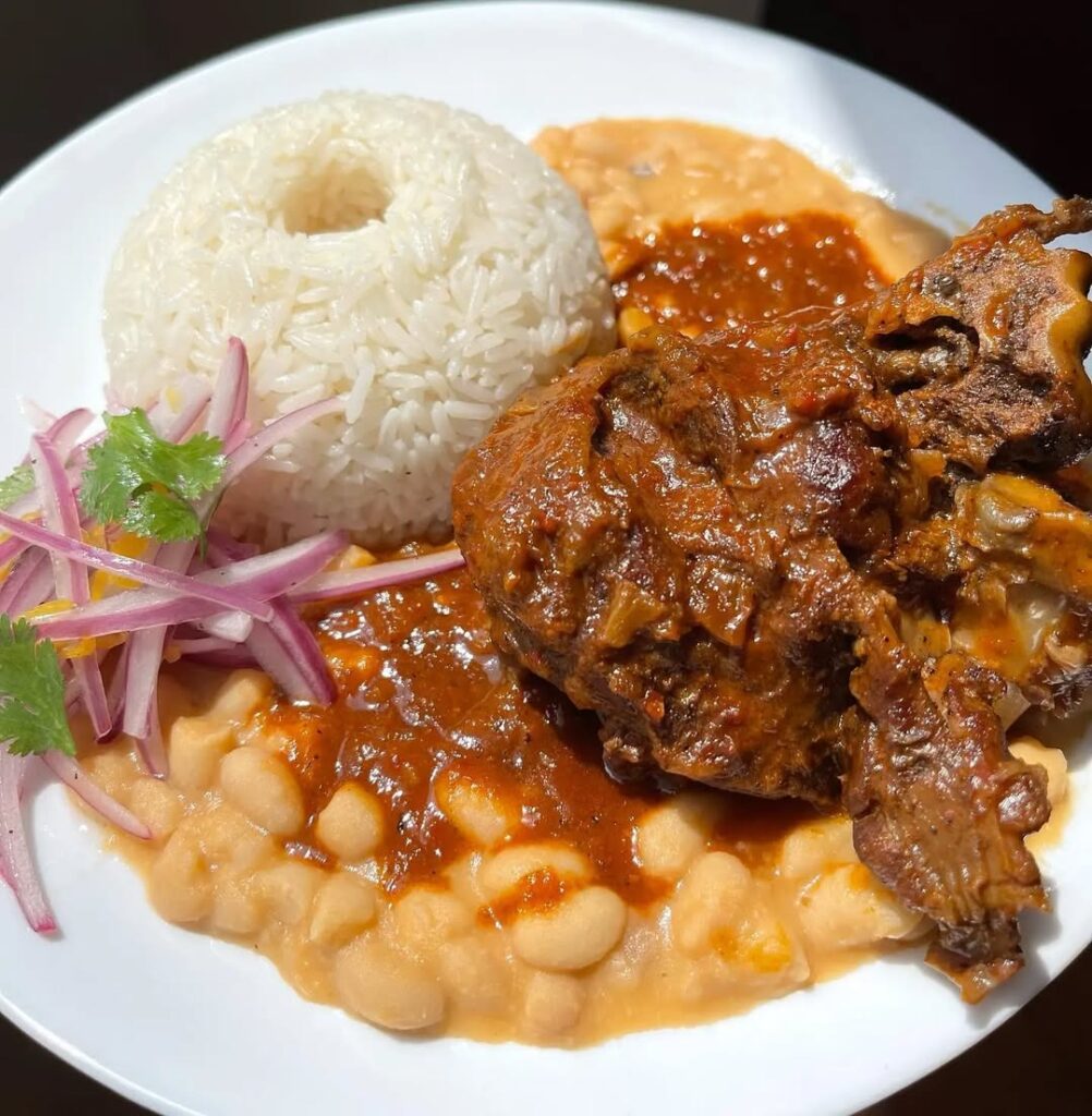 Seco de cabrito a la norteña acompañado de arroz, frejoles y cebolla encurtida, un guiso tradicional de Lambayeque y Piura que destaca por su sabor intenso y aroma a chicha de jora