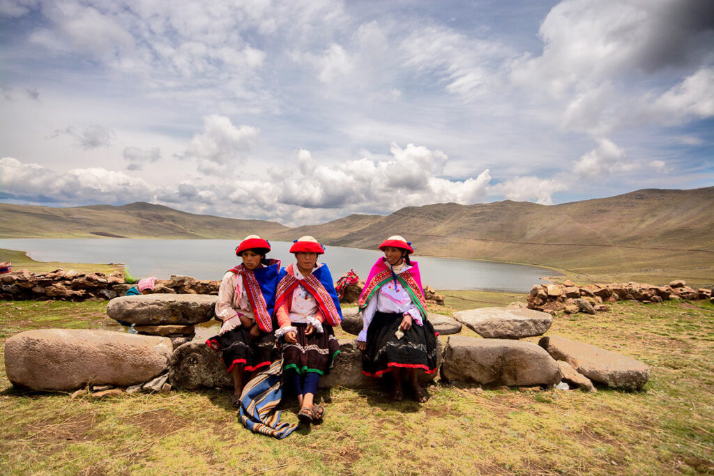 Comuneras andinas con vestimenta tradicional junto a la laguna de Qoricocha en Cusco, rodeadas de montañas y cielos despejados