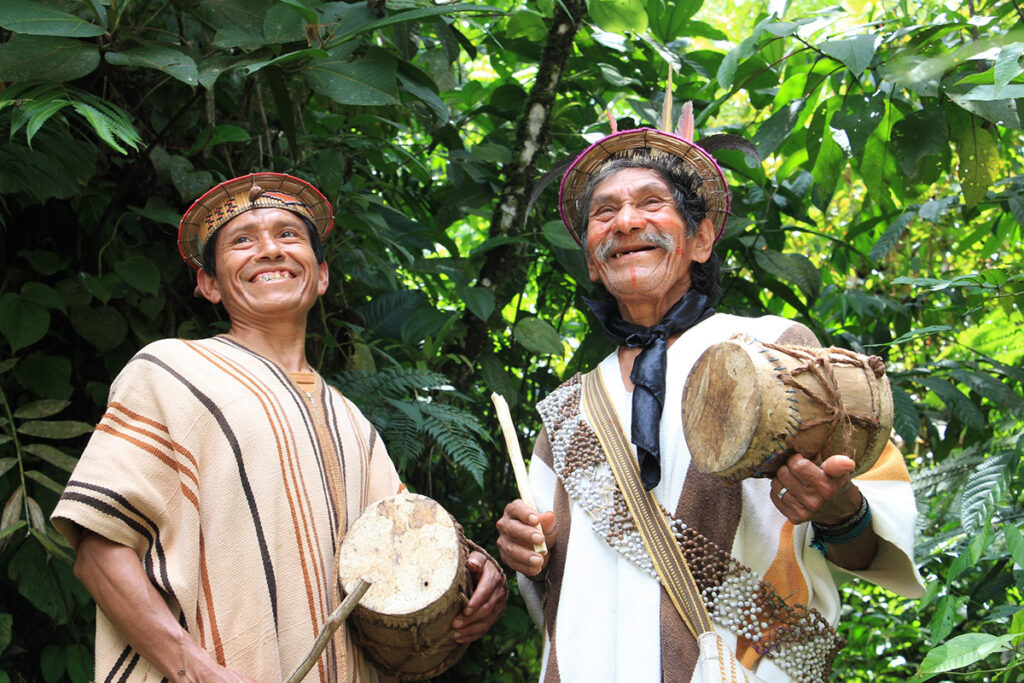 Dos hombres yánesha sonríen mientras tocan tambores tradicionales en medio de la selva central del Perú, símbolo de la alegría y vitalidad cultural del turismo sostenible comunitario