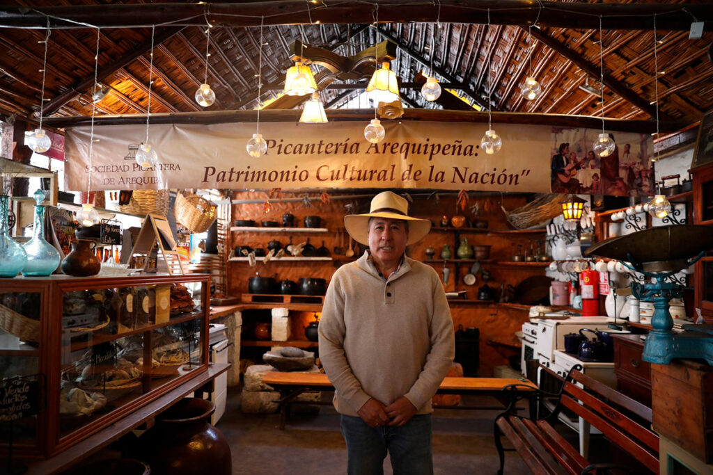 Interior de una picantería arequipeña tradicional con decoración rústica, estantes de utensilios antiguos y un hombre de sombrero posando junto a un cartel que la reconoce como Patrimonio Cultural de la Nación