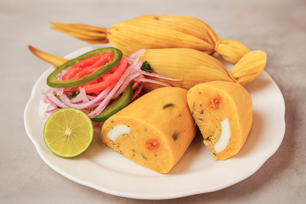Tamal cajamarquino servido con cebolla, tomate y limón sobre plato blanco, preparado con maíz morocho, manteca y ají colorado, típico de la sierra norte del Perú