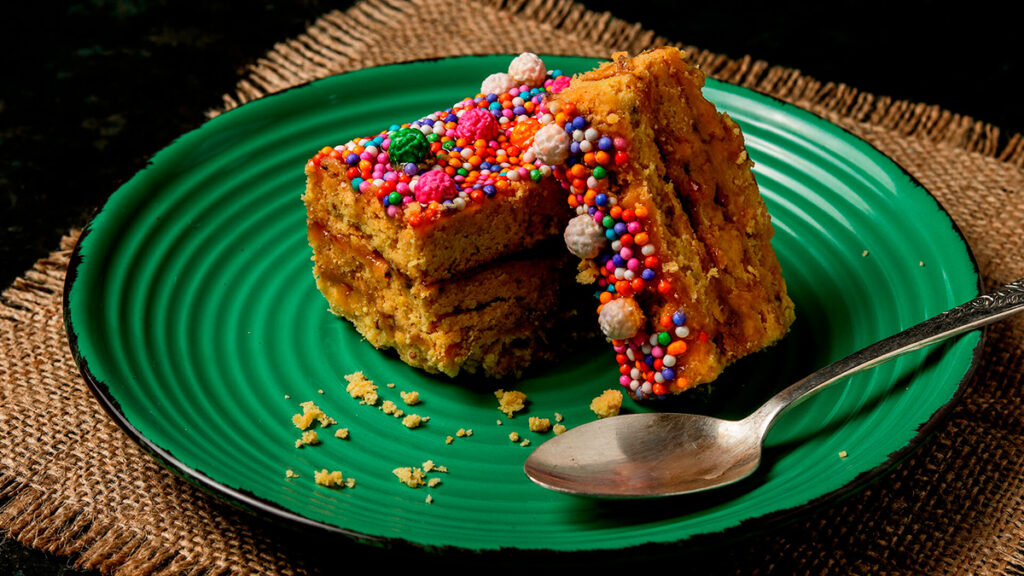 Porción de turrón de Doña Pepa decorado con grageas de colores, servido en un plato verde con cuchara metálica