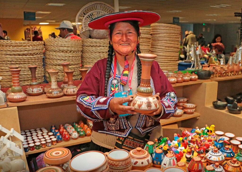 Artesana de Raqchi con sombrero tradicional sosteniendo una jarra decorada, rodeada de piezas de cerámica andina exhibidas en un puesto de mercado