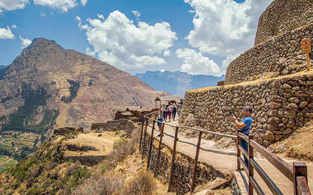 Sendero de acceso en Pisac con visitantes caminando junto a muros de piedra y montañas del Valle Sagrado al fondo
