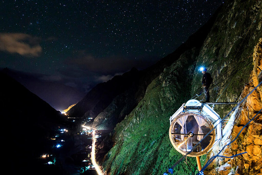 Cápsula del Skylodge iluminada de noche sobre la pared rocosa, con el cielo estrellado y el Valle Sagrado al fondo