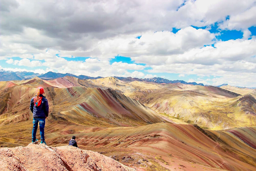 Visitante de pie frente a las laderas multicolores de la Montaña Palcoyo, con franjas rojizas, ocres y verdosas visibles desde un mirador alto