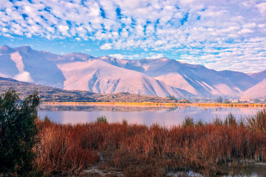 Vista panorámica de la laguna de Huacarpay con montañas del Valle Sur de Cusco reflejadas en sus aguas tranquilas, bajo un cielo despejado típico de la temporada seca