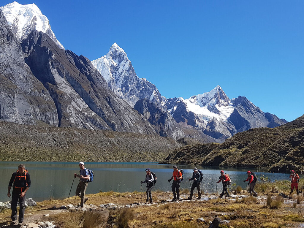 Grupo de trekkers caminando en altura en la Cordillera Huayhuash, con laguna altoandina y cumbres nevadas marcando el ritmo del recorrido